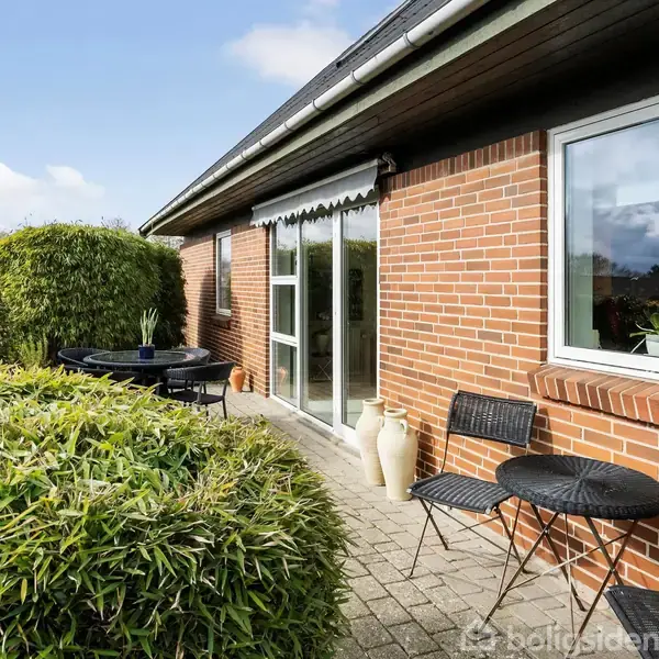 En rød murstenshusterrasse med havemøbler bestående af et sort bord og stole. To store krukker står ved den glasdør, der fører ind til huset. Omkring er grøn beplantning.