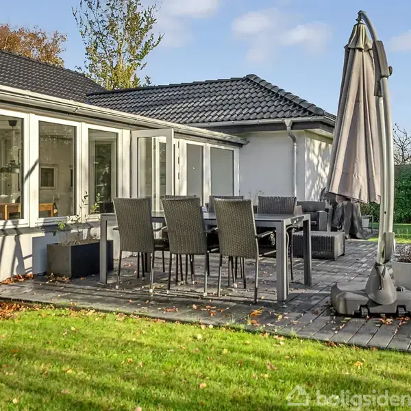 Terrassebord med seks stole står under en stor parasol på en træterrasse. Omkranset af en græsplæne, er terrassen tilknyttet et hvidt hus med store vinduer.