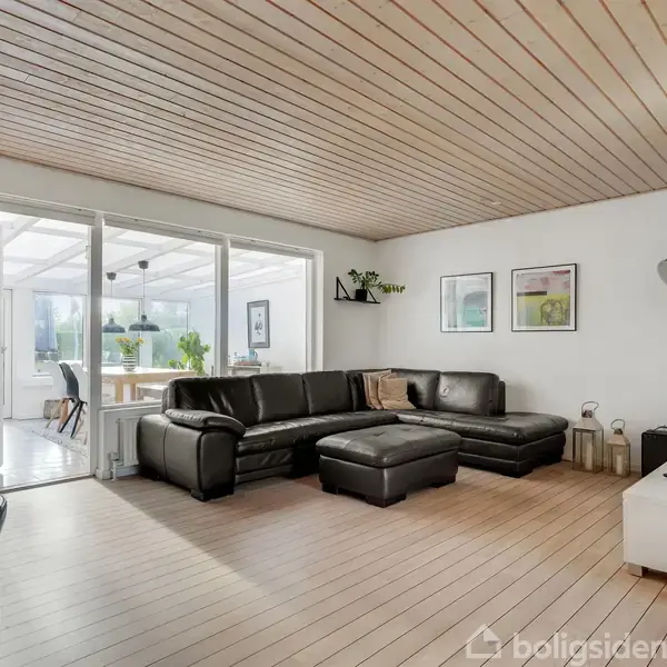 Sort læderhjørnesofa står i en stue med lyse trægulve og træloft. Ved siden af ses et hvidt tv-bord, og i baggrunden en glasdør til en lys udestue.