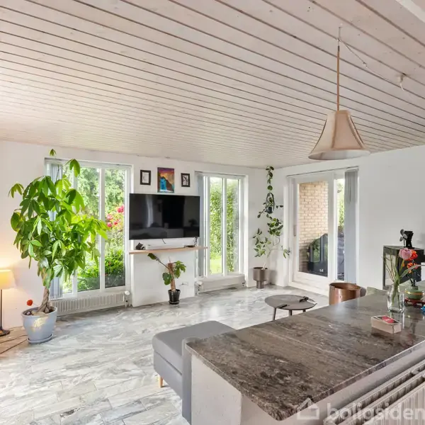 Stue med sofahjørne og fladskærms-TV på væggen, omgivet af grønne planter og store vinduer, der lukker naturligt lys ind. Køkkenø med pendellamper i forgrunden.