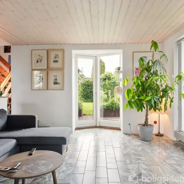 Sofa står i en lys stue ved siden af et rundt træbord. En stor plante ved franske døre giver udsigt til en have. Loftet er hvidt, og gulvet flisebelagt. Malerier hænger på væggen.