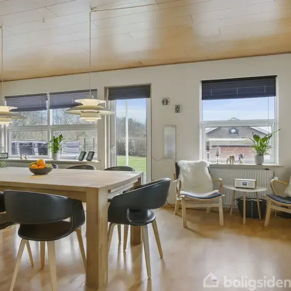 En moderne stue med en grå sofa omkring et træbord. Rummet har lænestol, barvogn og stort vindue, der slipper naturligt lys ind. Træloft og lyse trægulve skaber en lys atmosfære.