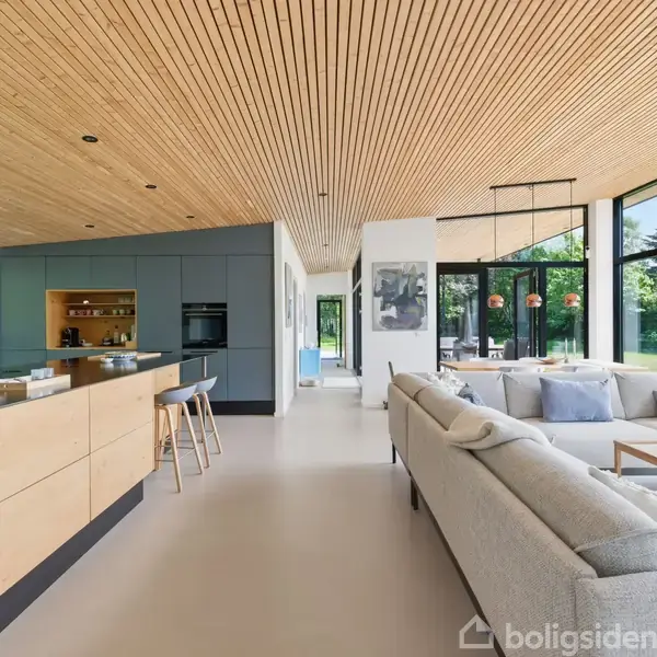 Køkken og stue er integreret i en moderne, lys bolig med trælofter, store vinduer og minimalistisk indretning. Køkkenøen har barstole, og stuen har en stor grå sofa.