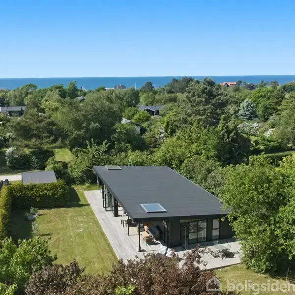 Hus med fladt tag står i solrigt haveområde omringet af træer; i baggrunden ses havet under en klar blå himmel.