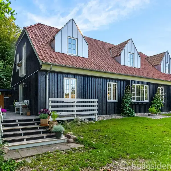 Sort træhus med rødt tegltag, hvide gavlvinduer og grønne klatreplanter, står i en frodig have med træer og terrasse med trapper.