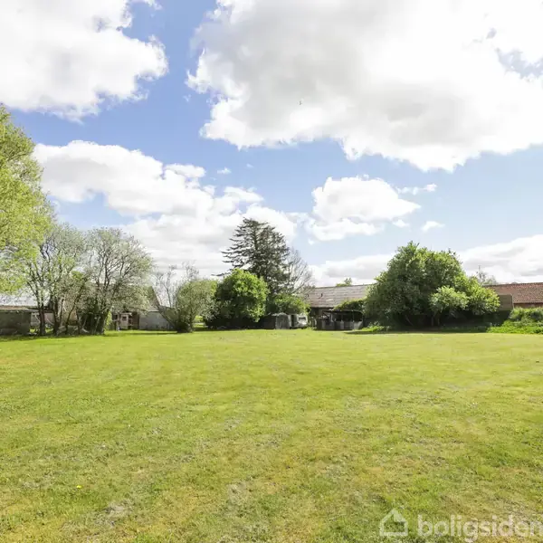 En stor, grøn græsplæne strækker sig fremad, omgivet af træer og buske under en delvist skyet blå himmel. I baggrunden ses en lav bygning og flere træer.