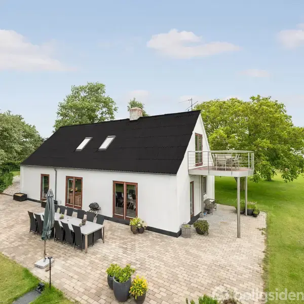 Et hvidt hus med sort tag står på en terrasse med havebord og stole. Omgivet af en grøn have med træer, mens en balkon stikker ud fra øverste etage.