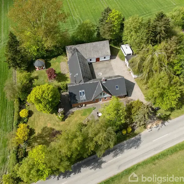 En stor ejendom omgivet af frodig vegetation ses fra oven. Bygningen ligger ved en vej, med gårdsplads og karport synlige. Omkringliggende marker strækker sig i baggrunden.