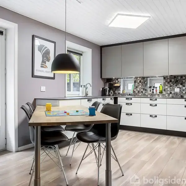 Køkkenbord med fire stole står i et moderne køkken. Skabe og skuffer i lyse farver. Væggen har mosaikfliser. Et vindue og glasdør fører ud til en træterrasse.