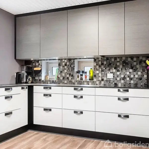 Køkken med grå og hvide skabe, arrangeret i en L-form, med en mosaikflisevæg som baggrund. Efterårsblad-dekoreret viskestykke hænger ved siden af en køkkenvask under et vindue med udsigt til grønne planter.