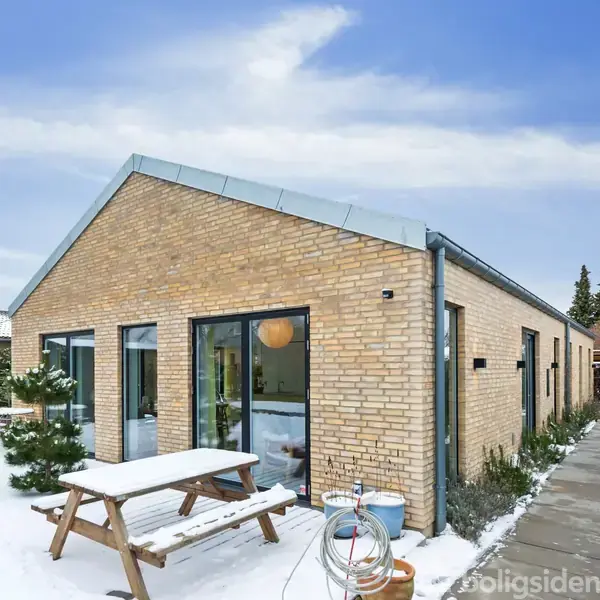 Et gult murstenshus med store vinduer står i et snedækket haveområde. En træbænk med et bord er placeret foran huset.