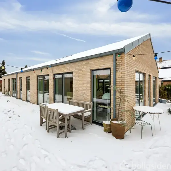 En moderne murstensbygning står dækket af sne. Udendørs er der havemøbler og plantekasser. Farverige lyskæder hænger over terrassen, hvilket skaber en stemningsfuld atmosfære om vinteren.