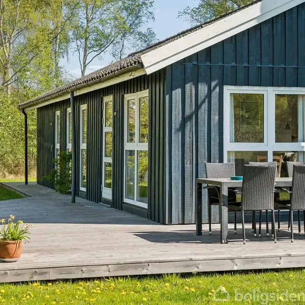 Blåt træhus med store vinduer står i solbelyst have. Terrasse med sorte stole og bord. Potteplante til venstre. Omgivet af grønne træer og græsplæne.