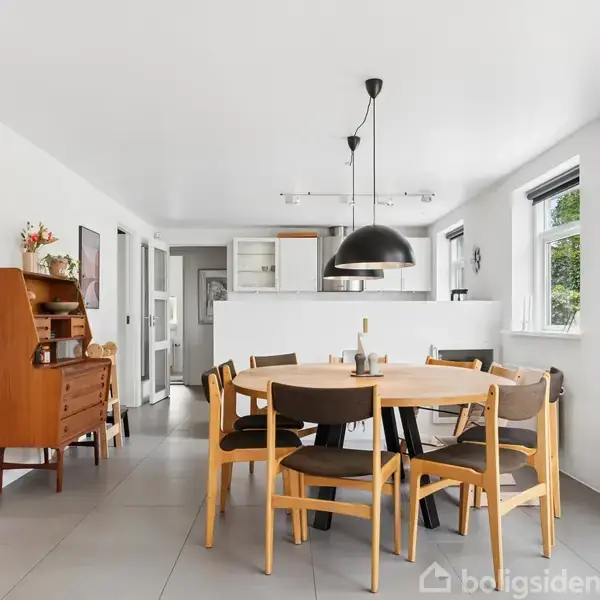 Et rundt spisebord med syv stole står i et lyst køkken-alrum. Over bordet hænger en sort pendellampe. Konteksten er minimalistisk med hvidmalede vægge og store vinduer.