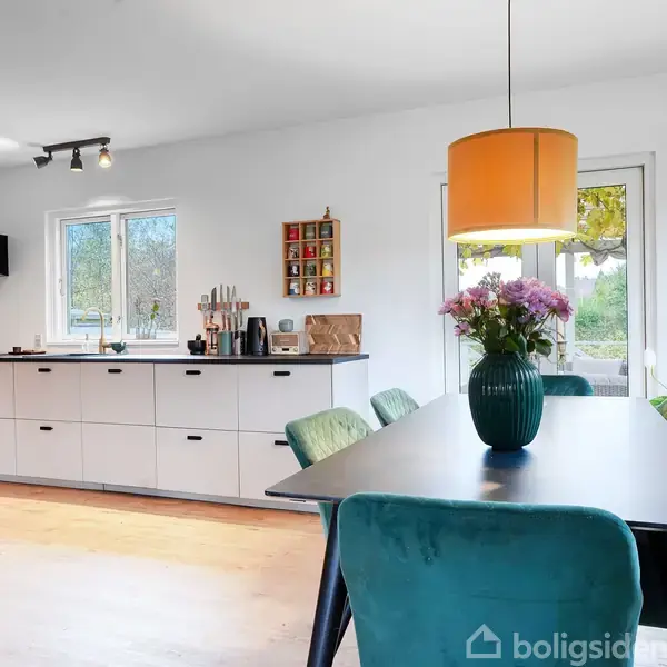 Et moderne køkken-alrum med et spisebord beklædt med turkise stole. En lampe hænger over bordet. Køkkenet har hvide skabe. En vase med blomster står på bordet.