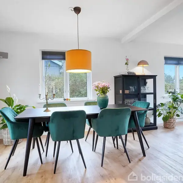 Spisebord omgivet af seks turkis stole står i en lys spisestue med trægulv og stor gul lampe. En sort skænk og planter dekorerer rummet. Plakat af Frida Kahlo ses.