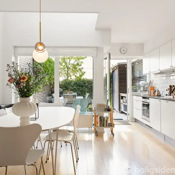 En moderne, lys spisestue-køkken kombination med et ovalt spisebord, hvide stole, vase med blomster, og hvidt køkkenskab. Store vinduer åbner op til en terrasse med grønne planter.