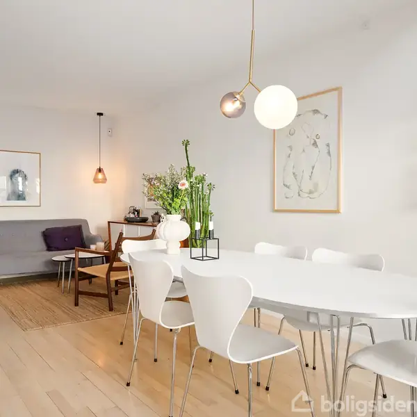 Spisebord med hvide stole står i en lys spisestue med trægulv. En sofa og lampe i baggrunden giver en hyggelig atmosfære. Væggene har kunstværker og en hylde med porcelæn.
