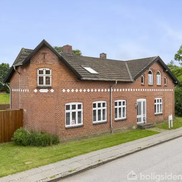 Et rødt murstenshus med hvide vinduer står ved en sti, omgivet af græs og en træhegn. Huset har en brun tagbeklædning med flere kviste og en solfanger.
