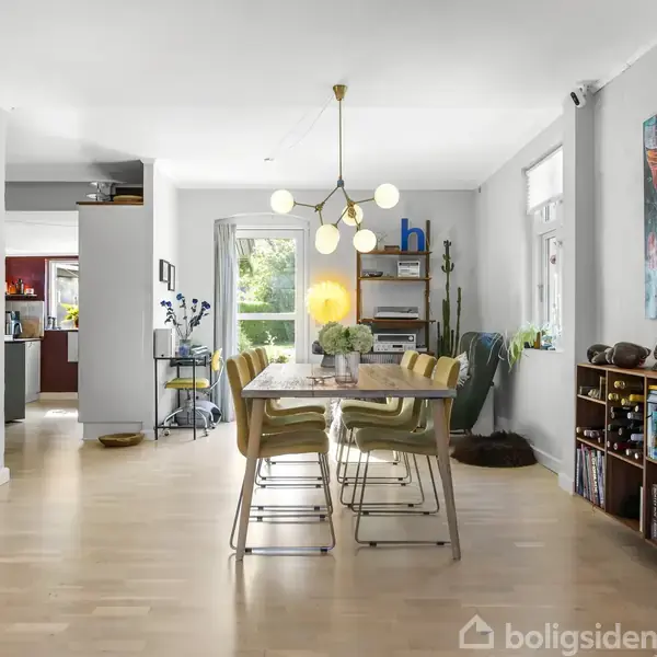Spisebord med syv stole er placeret i en lys spisestue. En moderne lysekrone hænger over bordet, og et farverigt maleri pryder væggen. Bogreol og lamper skaber hygge.