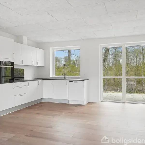 Køkken med hvide skabe og integrerede apparater i en lys og rummelig åben planløsning. Store vinduer og en havedør viser udsigt til grønne træer udenfor.