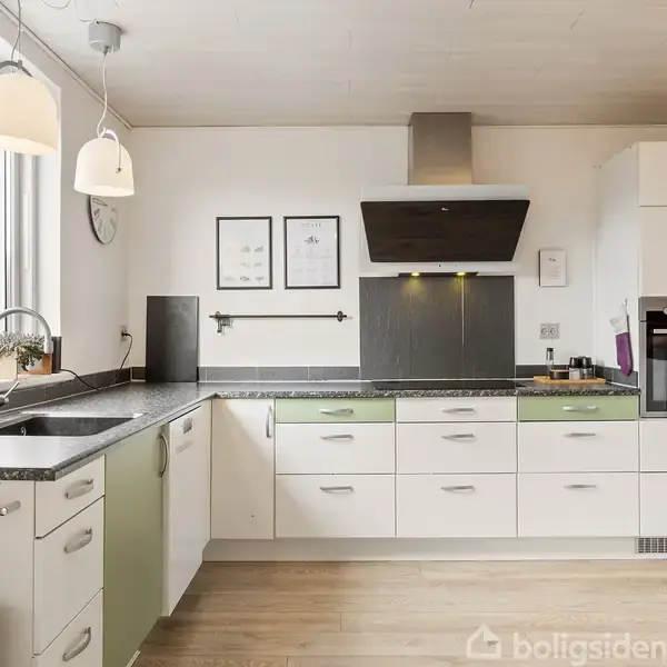 Et lyst køkken med hvide og grønne skabe, hvor der står en kaffemaskine på bordet. Væggene har indrammede billeder. Et stort vindue giver udsigt til en murstensbygning.