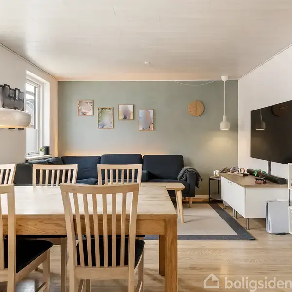 En spisestue med et træbord og stole står i centrum. Til højre ses en reol med dekorationer under et vægmonteret TV. En sofa og billeder pryder væggen i baggrunden.