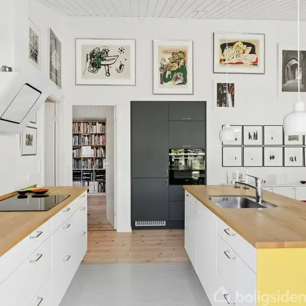 En moderne køkken med hvide skabe og træbordplader, herunder vask og kogeplade. Kunstværker hænger på væggen. En dør åbner ind til et rum med bogreoler.