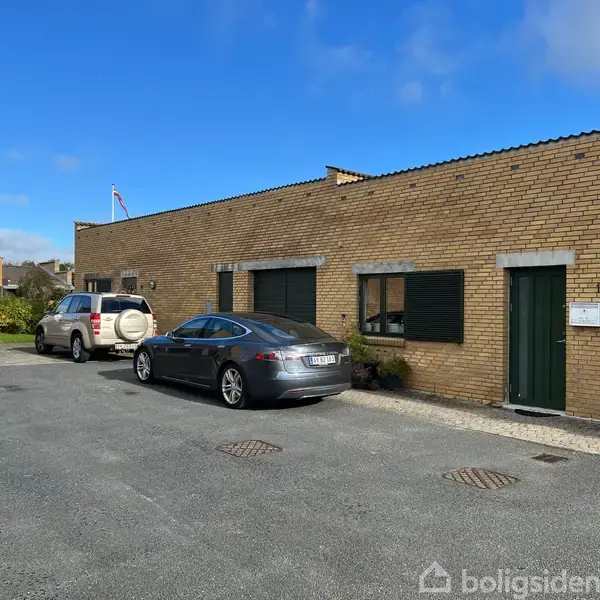 En grå bil holder parkeret foran en gul murstensbygning med grønne skodder. To andre biler er parkeret ved siden af på asfalten. Himlen er blå med nogle skyer.