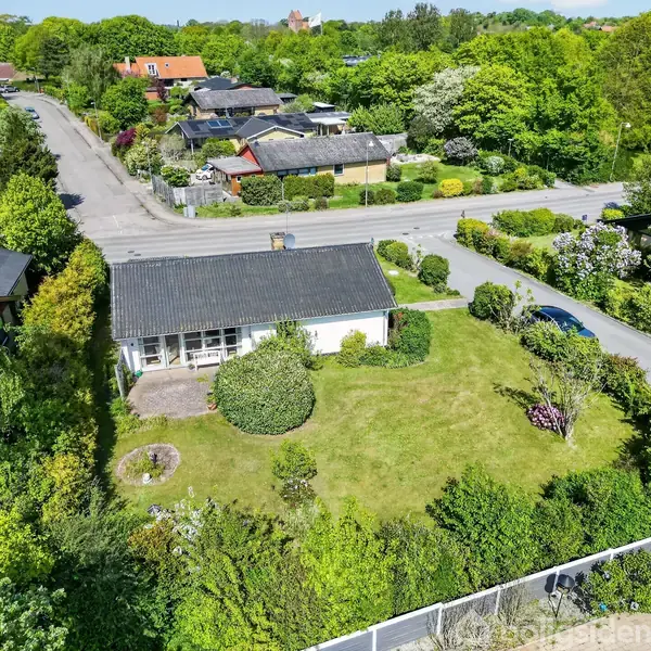 Et parcelhus med sort tag står omgivet af en velplejet have. Det er beliggende på en hjørnegrund i et grønt, frodigt boligområde med flere nærliggende villaer og veje.