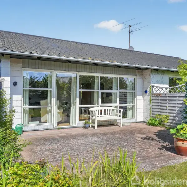Et enkelt hus med store glaspartier i facaden. En hvid bænk står på en terrasse omgivet af krukker med planter. Grønt buskads omkranser haven.