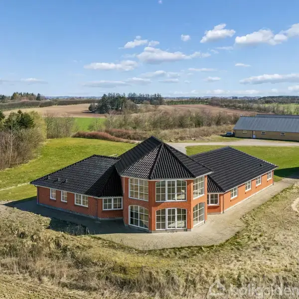 Et rødt murstenshus med sort tegltag står på en åben mark. Huset har store, buede vinduer og et rummeligt design. Omkringliggende marker og enkelte bygninger ses i baggrunden.