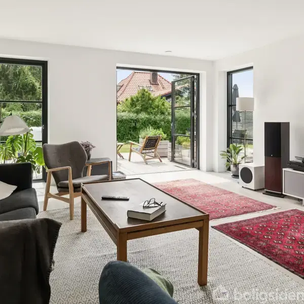 Stue med sofa og lænestol, hvor der står et sofabord med en bog. Store vinduer åbner ud til en terrasse med stole. Vægmonteret TV og røde tæpper på gulvet.
