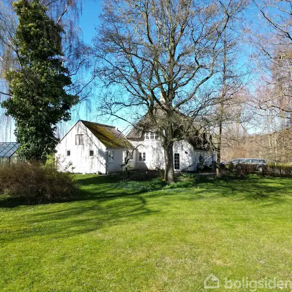 Hus omgivet af grøn have med store træer. I baggrunden ses en bil og en lav bygning. Skyfri blå himmel over landskabet.