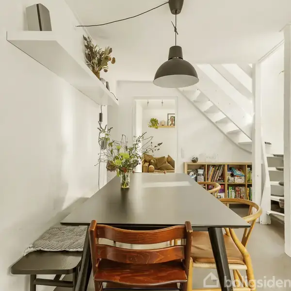 Et spisebord med en buket blomster står i et lyst rum. Til højre er der en reol med bøger og en trappe, der fører op. En sofa ses gennem døråbningen i baggrunden.