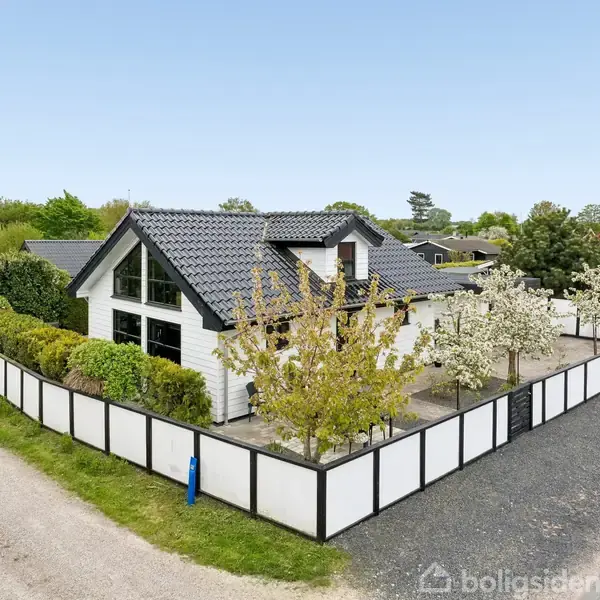 Hus med sort tag og store vinduer er omgivet af en lav hvid stakit og blomstrende træer. Beliggende ved en grusvej i et grønt, landligt område.