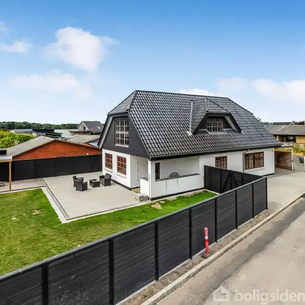 Et moderne hus med mørkt tegltag står stille. Omgivet af en hæk og fliser, med en græsplæne til venstre. En vej løber langs ejendommen.