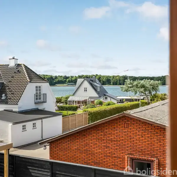 En hvid villa med stejlt tag er set fra oven, omgivet af grønne haver. I baggrunden ses en sø og en skov.