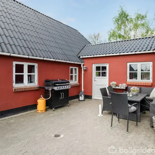 Rød murstensbygning har en åben gårdsplads med en grill til venstre og et sort spisebordssæt med seks stole til højre. Et lille bord har planter og en lysestage.