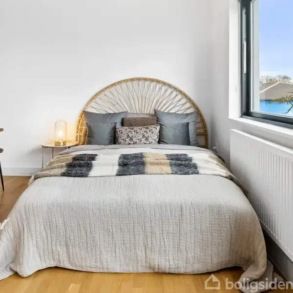 Seng med flettet hovedgærde; placeret mod en hvid væg. På sengetæppet ligger en ternet plaid. Til venstre står et lille bord med stol og lampe. Vindue til højre.