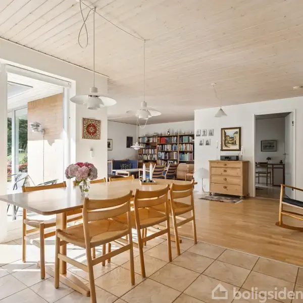 Spisebord med otte træstole, dekoreret med blomster, står i et lyst rum med åbne døre til en terrasse. Reoler med bøger og skænk ses i baggrunden.