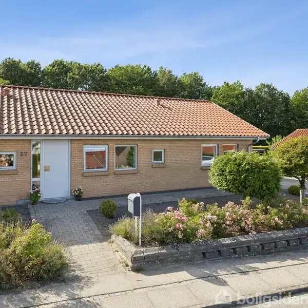 Et enfamiliehus med rødt tegltag og beige murstensfacade. Huset har en hvid hoveddør og flere vinduer. Forhaven er velholdt med blomsterbede og buske. Nummer 37 er synligt på væggen.