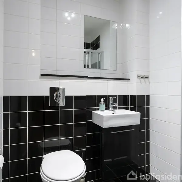 Håndvask med vandhane ved siden af en bruseniche. Brusenichen har sort/hvide fliser i striber. Badeværelsesinteriøret er moderne med skarpe linjer. Ingen tekst er synlig.