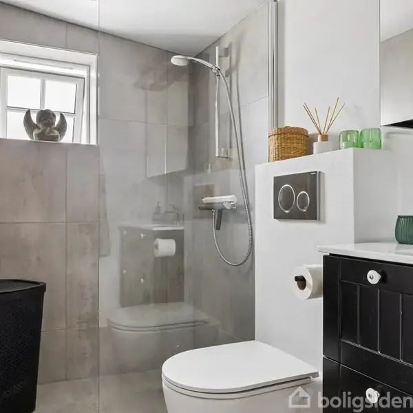 Badeværelse med væghængt toiletskål ved siden af en glasbrusekabine. Et vindue over bruseren bidrager med lys, og en sort skuffeenhed står ved siden af. Der findes ingen synlig tekst.