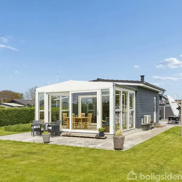 Et moderne sommerhus med store vinduer står på en terrasse omringet af en velplejet græsplæne. Et spisebord med stole er synligt gennem vinduerne.