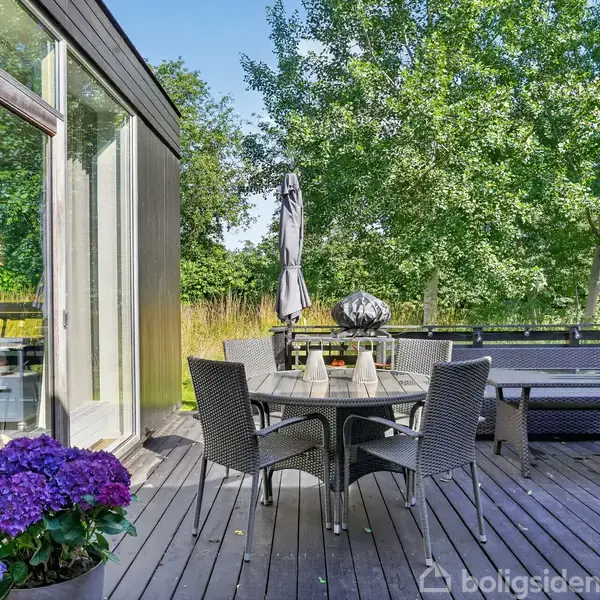 Terrasse med rundt bord og seks stole arrangeret omkring det. Blomsterkrukke med lilla blomster står i hjørnet. Parasol og sofa i baggrunden omgivet af grønne træer.