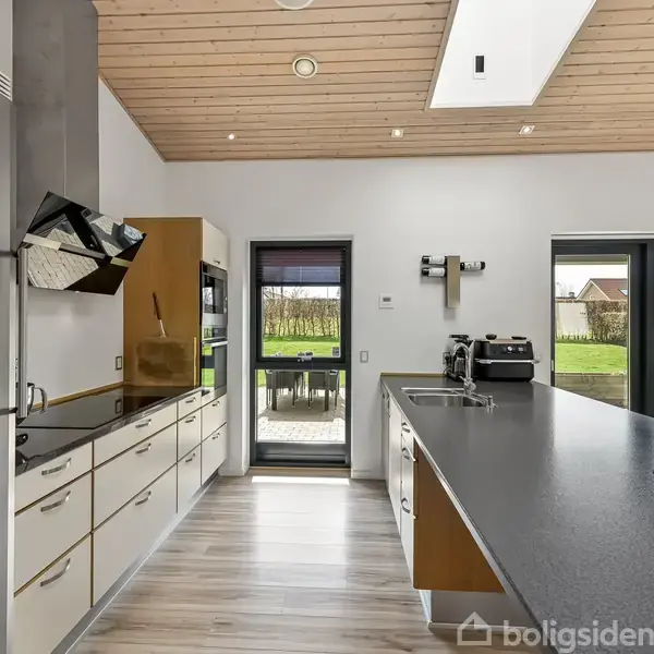 Et moderne køkken med højskabe og indbyggede apparater ved siden af en stor køkkenø med vask, overfor en glasdør og vinduer med udsigt til en grøn have.