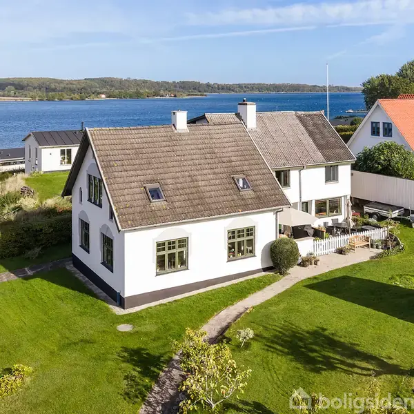 Hus med hvid facade og skifertag står stille i have, med terrasse foran. Det ligger ved kysten, med udsigt over havet og naboer tæt ved siden.