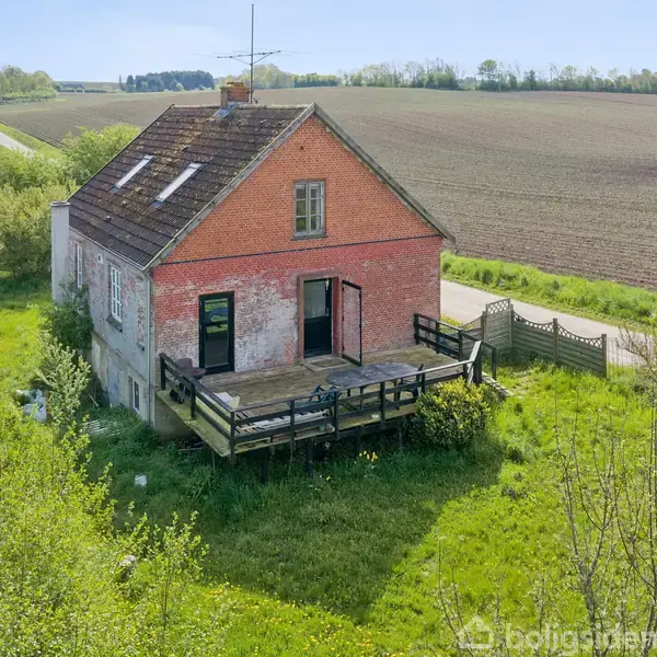 Et ældre hus med rød murstensfacade og stor træterrasse står stille på en grøn, tilgroet grund omgivet af åbne marker og en vej i det fjerne.
