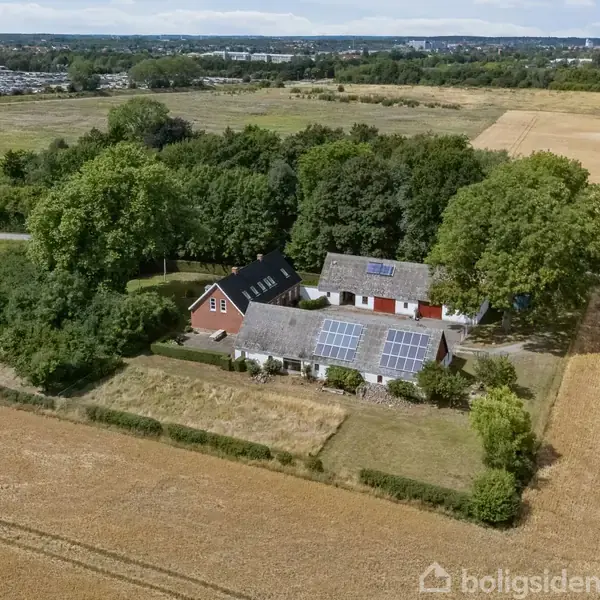 Et landhus med solpaneler på taget står omgivet af træer og marker. Baggrunden viser en landlig landskabshorisont med flere åbne marker.